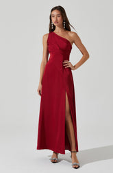 A woman is wearing a long, elegant, strapless red dress. The dress features a gathered, ruched bodice on one side and a high slit on the front left side, revealing her leg. She accessorizes with large, dangling earrings and open-toed heels with ankle straps.